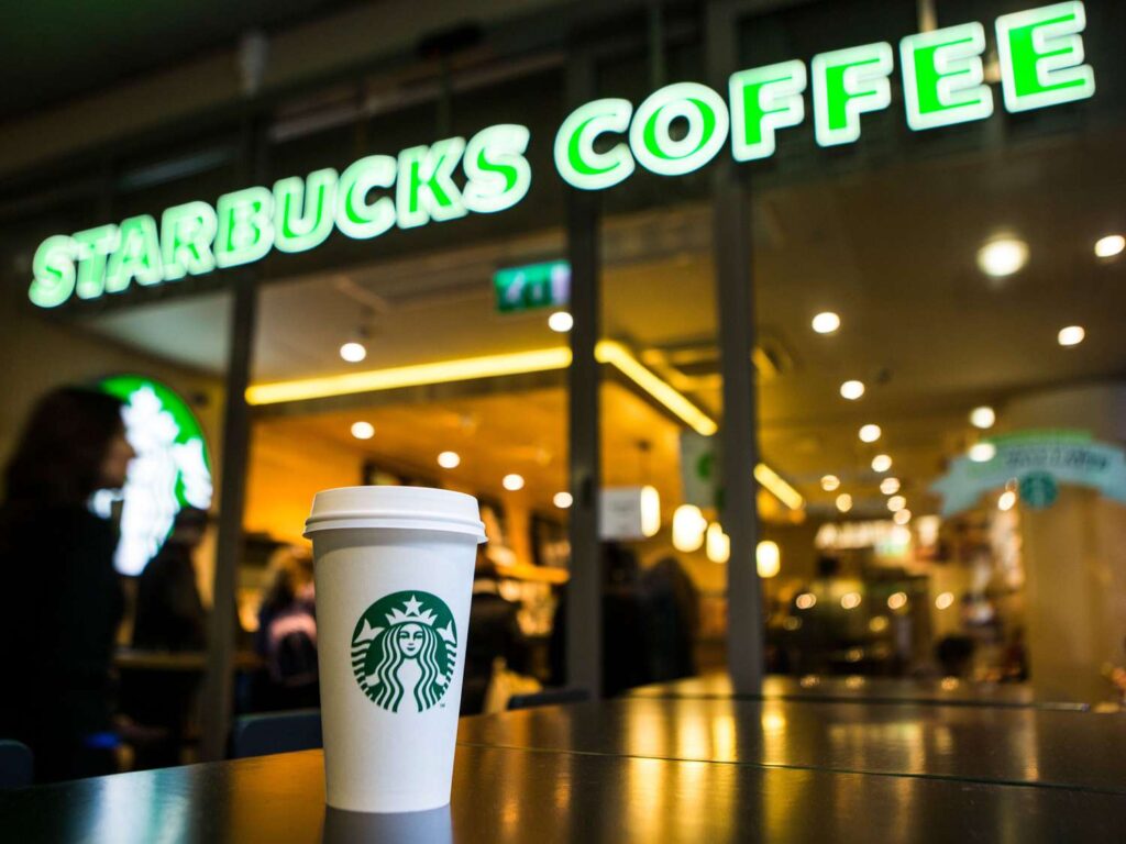 Starbucks coffee cup in foreground with Starbucks Coffee sign and blurred café interior, illustrating customer loyalty and personalized rewards.